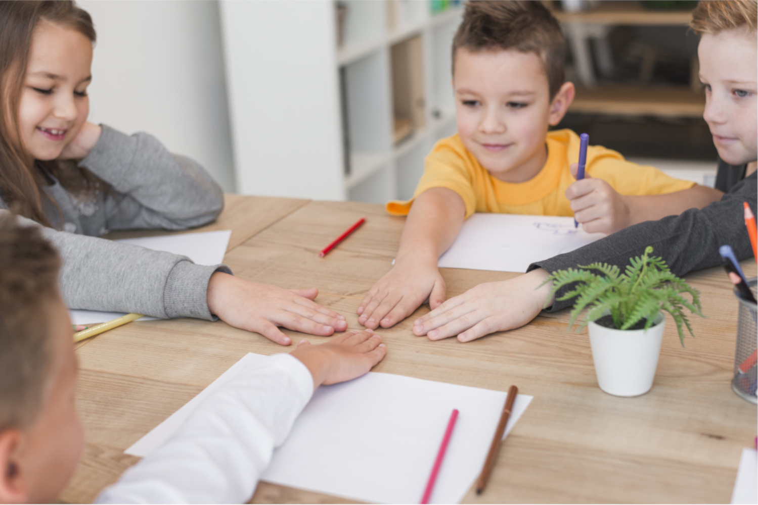 Ci sono quattro bambini seduti intorno ad un tavolo con le mani unite al centro, tre sono maschi ed una femmina. Stanno iniziando a scrivere e disegnare insieme, perché hanno tutti pennarelli e foglio di fronte a loro